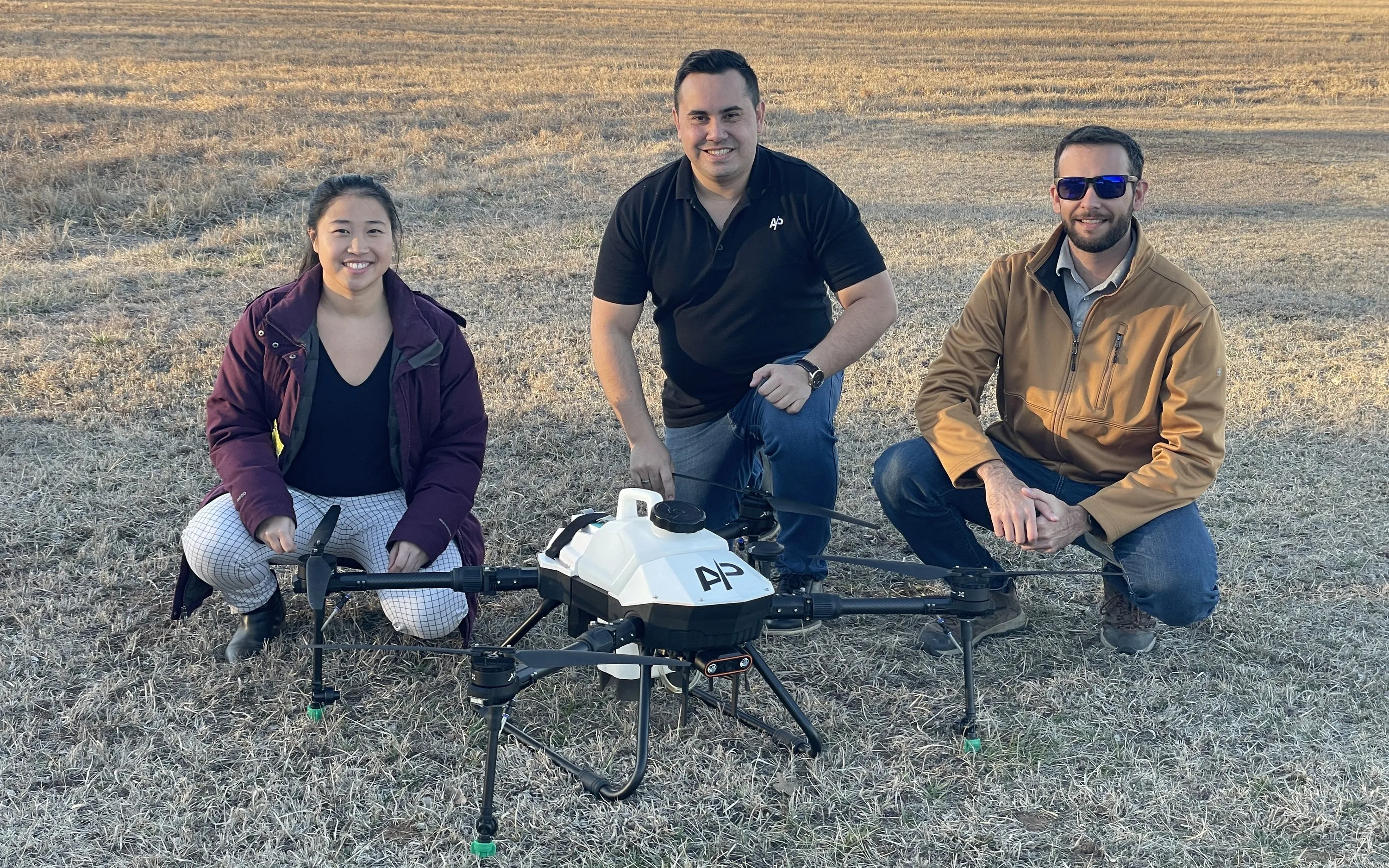 Three American Prime team members crouched beside an AP agricultural spray drone on dry winter grass